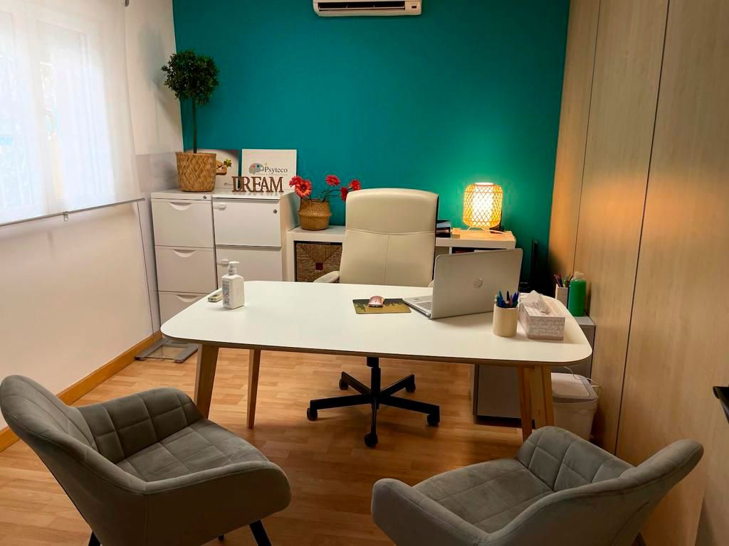 Imagen de despacho con mesa y silla de color blanco, dos butacas en frente de color gris y pared del fondo azul cobalto corporativo